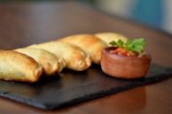 A row of golden-brown baked empanadas is arranged on a dark slate serving board, accompanied by a small clay bowl filled with a colorful salsa garnished with a sprig of parsley.