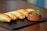A row of golden-brown baked empanadas is arranged on a dark slate serving board, accompanied by a small clay bowl filled with a colorful salsa garnished with a sprig of parsley.