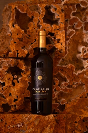 A bottle of Chaglasian Master Blend wine is positioned against a textured, rust-colored rocky background that has intricate patterns and a natural, rugged appearance. The wine bottle features a dark label with elegant golden text and design, topped with a gold foil cap.