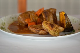 A plate of food featuring roasted potato wedges, slices of meat in a rich brown sauce, and a piece of roasted squash, garnished with herbs. The dish is served on a white plate against a blurred background.