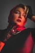 A person wearing a red headscarf and black lace gloves poses with an elegant expression, featuring bold red lipstick. The lighting creates dramatic shadows, highlighting the glossy texture of the scarf and the intricacy of the lace gloves.