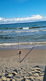 A fishing rod is planted upright in the sandy shore near the waterline of a beach. The waves of the ocean are gently rolling in under a clear blue sky with some clouds in the distance.