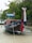 A traditional wooden long-tail boat rests on a sandy beach, adorned with colorful ribbons. The boat displays writing on its side and a flag at the stern. It is bordered by dense greenery, creating a serene and natural setting.