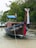 A traditional wooden long-tail boat rests on a sandy beach, adorned with colorful ribbons. The boat displays writing on its side and a flag at the stern. It is bordered by dense greenery, creating a serene and natural setting.
