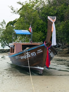 A traditional wooden long-tail boat rests on a sandy beach, adorned with colorful ribbons. The boat displays writing on its side and a flag at the stern. It is bordered by dense greenery, creating a serene and natural setting.