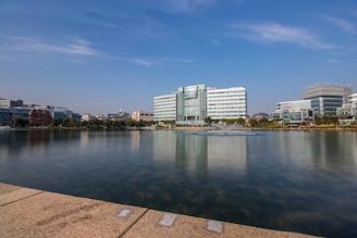 A waterfront view featuring a series of modern office buildings lining the edge of a tranquil body of water. In the foreground, a stone path borders the calm lake which reflects the blue sky and the buildings. A central fountain is visible in the water, surrounded by various trees and sparse vegetation.