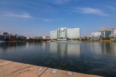 A waterfront view featuring a series of modern office buildings lining the edge of a tranquil body of water. In the foreground, a stone path borders the calm lake which reflects the blue sky and the buildings. A central fountain is visible in the water, surrounded by various trees and sparse vegetation.