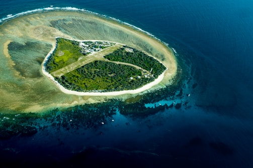 An aerial view of a small island surrounded by deep blue ocean waters. The island is covered with dense green vegetation and appears to have some man-made structures and a narrow dirt path traversing across it. The surrounding shallow waters exhibit a lighter shade of blue with visible sandbars and coral formations.