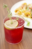 A bright red beverage served in a textured glass garnished with a slice of lemon and a sprig of herbs, with a plate of food blurred in the background, placed on a wooden surface.