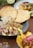 A plate with diced raw fish mixed with onions, herbs, and sprouts is garnished with lime slices and accompanied by round, crispy corn tortillas. In the background, another dish topped with grilled vegetables can be seen, and a small bowl containing tortilla chips with a meat and onion mixture is in the foreground.