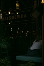 A dimly lit indoor scene features a partially visible person sitting near a window. The word 'MALQUERIDA' is prominently displayed on a sign. Several decorative lanterns hang from above, casting warm glows, while potted plants add greenery in the foreground.