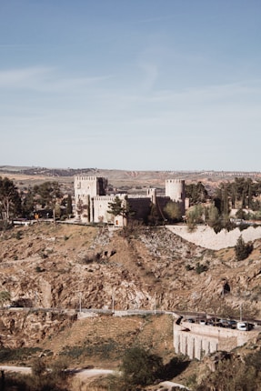 A historic castle with fortified stone walls and towers sits atop a rugged hill surrounded by lush greenery. The landscape extends to distant rolling fields under a clear blue sky, creating a picturesque scene.