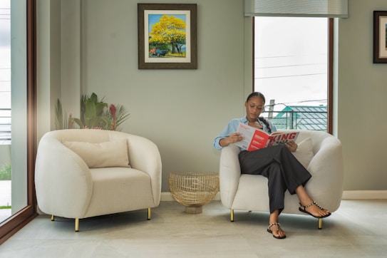 A person is seated in a modern living room with minimalist furniture. They are reading a magazine while sitting on a cushy, white armchair. Another similar armchair is adjacent to it, separated by a small, decorative wicker basket. The walls are painted a soothing green, and there are two framed artworks on them. The room has large windows, providing ample natural light.