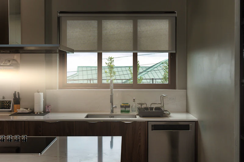 Modern kitchen with dark cabinetry, white countertop, stainless steel sink and window with Roman shade