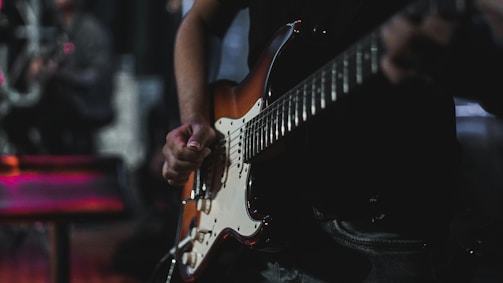 A person playing an electric guitar, focusing on the hand strumming the strings. The guitar is a classic sunburst design and the scene is dimly lit, suggesting a performance or practice environment. The blurry background indicates a stage or music venue setting.