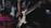 A person playing an electric guitar, focusing on the hand strumming the strings. The guitar is a classic sunburst design and the scene is dimly lit, suggesting a performance or practice environment. The blurry background indicates a stage or music venue setting.