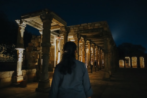 An individual is standing in front of an ancient stone structure illuminated by lights at night. The architecture features columns and arches, conveying a historical or archaeological site ambiance.