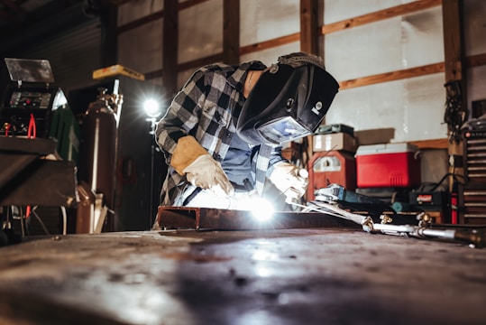 A person wearing protective gear and a welding helmet is engaged in welding activities, surrounded by various tools and equipment in a workshop environment. The bright light from the welding process illuminates the dark space, highlighting the focus and intensity of the work. The setting includes items such as gas tanks, a red cooler, and assorted tools on shelves.