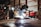 A person wearing protective gear and a welding helmet is engaged in welding activities, surrounded by various tools and equipment in a workshop environment. The bright light from the welding process illuminates the dark space, highlighting the focus and intensity of the work. The setting includes items such as gas tanks, a red cooler, and assorted tools on shelves.