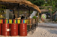 A tropical bar with a thatched roof is situated on a sandy surface. The bar has colorful painted barrels lined up in front, along with a series of tall chairs. Behind the bar, various bottles are visible on a shelf, and a chalkboard menu is also present. In the background, there is a sign that reads 'DREAM Resort', surrounded by lush greenery and a stone pathway leading upwards. A stone with a map engraved on it is also nearby.