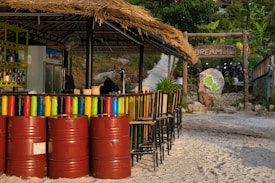A tropical bar with a thatched roof is situated on a sandy surface. The bar has colorful painted barrels lined up in front, along with a series of tall chairs. Behind the bar, various bottles are visible on a shelf, and a chalkboard menu is also present. In the background, there is a sign that reads 'DREAM Resort', surrounded by lush greenery and a stone pathway leading upwards. A stone with a map engraved on it is also nearby.