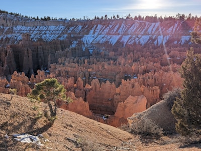 A breathtaking view of a canyon filled with uniquely shaped rock formations, known as hoodoos, which are illuminated by sunlight. Snow patches highlight the layered rock patterns while evergreen trees are scattered across the landscape.