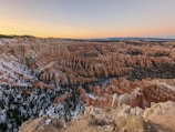 A breathtaking view of a vast canyon landscape with unique rock formations known as hoodoos, characterized by their tall, thin spires. The colors range from orange to red, with patches of green vegetation and some snow covering. The sky is clear with a gradient from light blue to a soft peach tint as the sun sets.