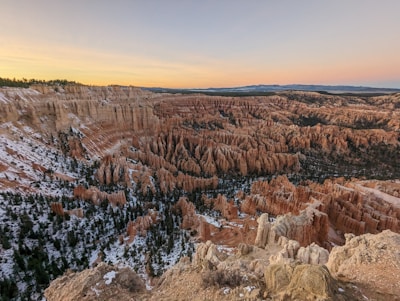 A breathtaking view of a vast canyon landscape with unique rock formations known as hoodoos, characterized by their tall, thin spires. The colors range from orange to red, with patches of green vegetation and some snow covering. The sky is clear with a gradient from light blue to a soft peach tint as the sun sets.