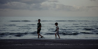 Two children are walking along the shoreline on a beach with gentle waves in the evening or early morning light. The sky is overcast with clouds, adding a moody atmosphere to the scene.