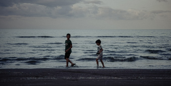 Two children are walking along the shoreline on a beach with gentle waves in the evening or early morning light. The sky is overcast with clouds, adding a moody atmosphere to the scene.