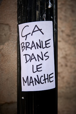 A handwritten sign on a white piece of paper is taped to a black pole. The text is in French, saying 'ÇA BRANLE DANS LE MANCHE.' The surrounding area is slightly blurred, giving focus to the sign.