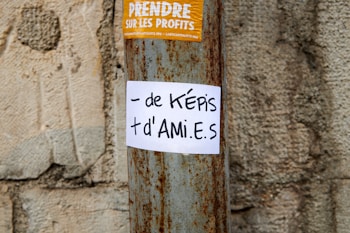 A worn concrete wall background is contrasted by a rusted metal pole. Attached to the pole is a white paper with handwritten black text that reads '- de képis + d'ami.e.s'. Above it, there is a yellow sticker with white text in French about taking profits.