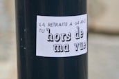A black pole with a white sticker that has French text. The text reads 'LA RETRAITE À 64 ANS TU hors de ma vue' which translates to 'The pension at 64 get out of my sight.'