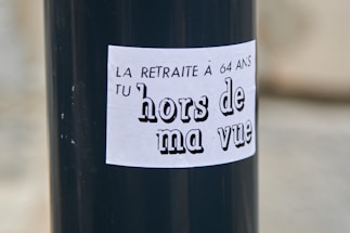 A black pole with a white sticker that has French text. The text reads 'LA RETRAITE À 64 ANS TU hors de ma vue' which translates to 'The pension at 64 get out of my sight.'