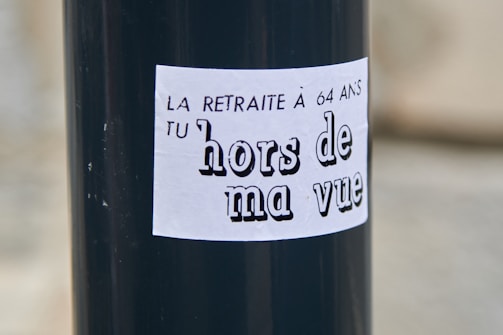 A black pole with a white sticker that has French text. The text reads 'LA RETRAITE À 64 ANS TU hors de ma vue' which translates to 'The pension at 64 get out of my sight.'