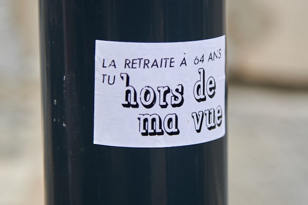 A black pole with a white sticker that has French text. The text reads 'LA RETRAITE À 64 ANS TU hors de ma vue' which translates to 'The pension at 64 get out of my sight.'