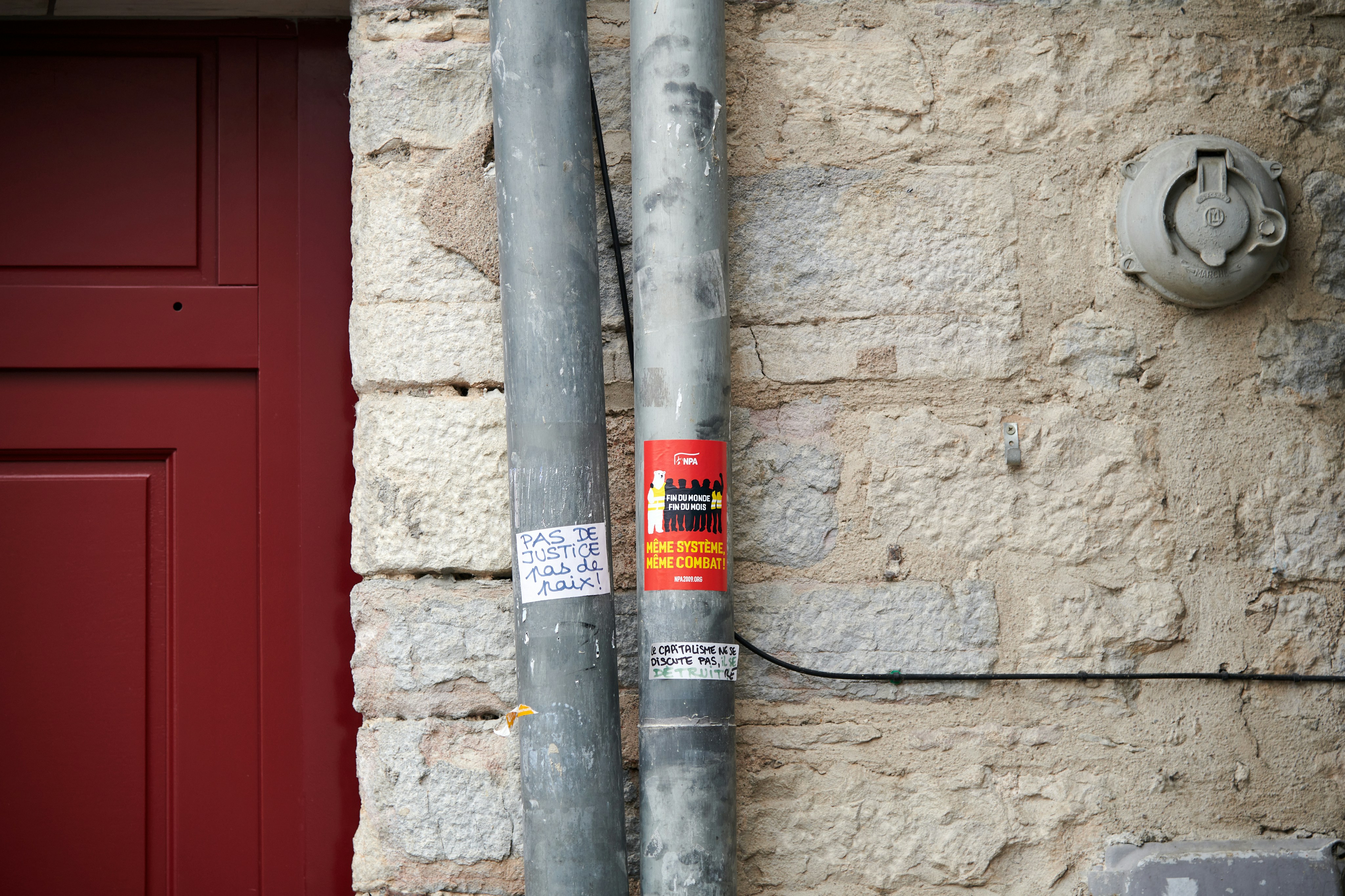A close-up of a beige stone wall with two vertical metal pipes secured to it. One of the pipes has a red sticker with text and images, and below it, there is a small handwritten note on white paper. To the left, part of a red door frame is visible. An electrical fixture is mounted on the wall to the right.