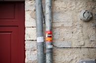 A close-up of a beige stone wall with two vertical metal pipes secured to it. One of the pipes has a red sticker with text and images, and below it, there is a small handwritten note on white paper. To the left, part of a red door frame is visible. An electrical fixture is mounted on the wall to the right.