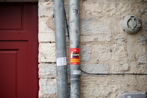 A close-up of a beige stone wall with two vertical metal pipes secured to it. One of the pipes has a red sticker with text and images, and below it, there is a small handwritten note on white paper. To the left, part of a red door frame is visible. An electrical fixture is mounted on the wall to the right.