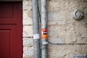 A close-up of a beige stone wall with two vertical metal pipes secured to it. One of the pipes has a red sticker with text and images, and below it, there is a small handwritten note on white paper. To the left, part of a red door frame is visible. An electrical fixture is mounted on the wall to the right.