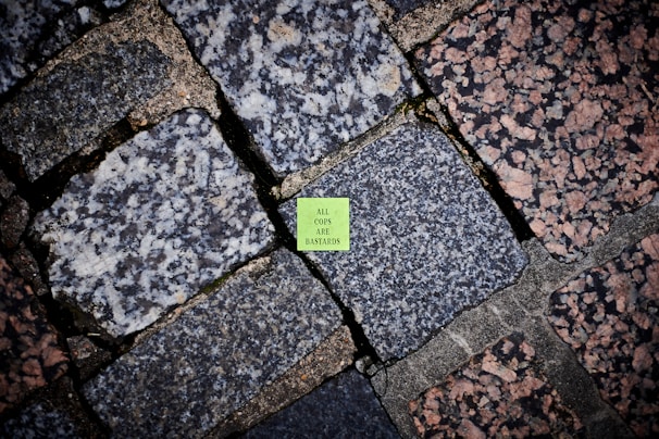 A vivid green sticker with text is placed on an outdoor stone pavement, surrounded by a variety of large, rectangular cobblestones with a mix of gray and pink tones. The stones have different textures, adding visual diversity to the ground surface.