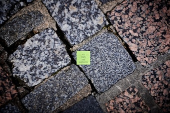 A vivid green sticker with text is placed on an outdoor stone pavement, surrounded by a variety of large, rectangular cobblestones with a mix of gray and pink tones. The stones have different textures, adding visual diversity to the ground surface.