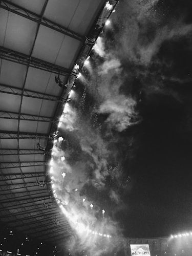 A black and white image of a stadium interior with a partial view of the roof and a display screen. Spotlights and smoke are visible, creating a dynamic atmosphere.
