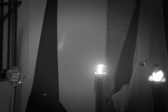 A figure dressed in a pointed hood and robe holds a lit candle. The background is dimly lit, creating a somber and mysterious atmosphere.
