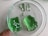 A close-up view of a petri dish containing two chunks of translucent green gelatin-like material with some white spots inside one of them. The petri dish is being held by a finger visible at the edge.