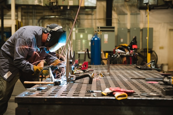 A worker wearing protective gear is engaged in welding on a large metal table. Sparks fly from the welding activity, illuminating the area around him. The workshop environment is industrial, with various tools and equipment visible in the background.