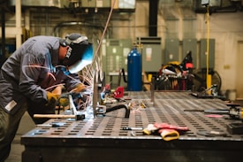 A worker wearing protective gear is engaged in welding on a large metal table. Sparks fly from the welding activity, illuminating the area around him. The workshop environment is industrial, with various tools and equipment visible in the background.