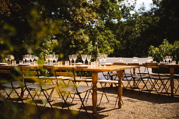 Wooden tables set with wine glasses on gravel under green trees