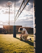 A football player is kneeling on the field near the goal post, holding his helmet. The scene is set during sunset with a dramatic sky. Stadium lights are visible, and there are buildings in the background. The grass is green and well-maintained.