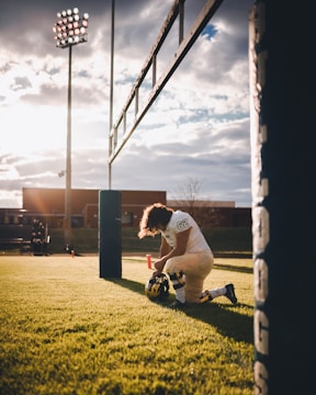 A football player is kneeling on the field near the goal post, holding his helmet. The scene is set during sunset with a dramatic sky. Stadium lights are visible, and there are buildings in the background. The grass is green and well-maintained.